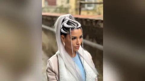 Woman with silver braided high ponytail and light blue outfit posing outdoors in soft natural light.