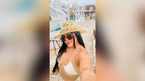 Smiling woman in white bikini and straw hat posing at a sunny outdoor resort with white statues.