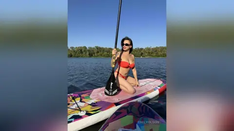 Woman in red bikini sitting on a colorful paddleboard on a sunny lake during a summer water sport activity.