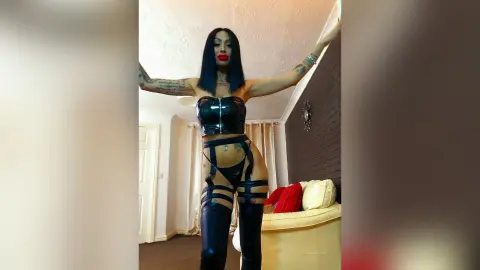 DeeaAnderson poses in black latex lingerie and thigh-high stockings, standing with her arms raised against a dark wall.