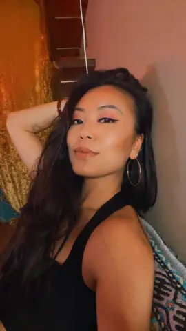 Serve_Cherry poses with one hand in her long dark hair, wearing a black tank top and gold hoop earrings.