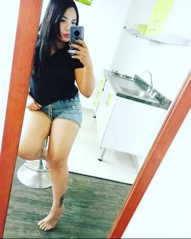 Young woman in black t-shirt and denim shorts taking a mirror selfie in a bright modern kitchen.