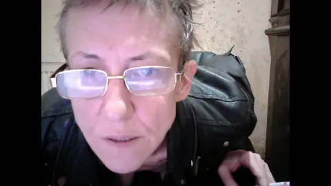 Close-up portrait of a woman with short hair wearing glasses and a black leather jacket in a dimly lit room.