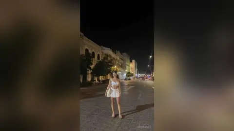 Young woman in a white summer outfit standing on a cobblestone street at night under city lights.