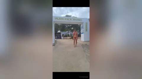 Woman in a red bikini standing under the Playa Cala Bassa entrance sign on a sunny beach day.