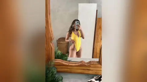 Woman in a bright yellow one-piece swimsuit taking a mirror selfie in a minimalist modern room.