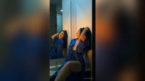 Elegant woman in a blue satin robe posing in a mirror with warm, moody lighting.