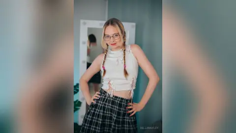Smiling young woman with blonde braids wearing glasses, a white crop top, and plaid skirt in a bright room.