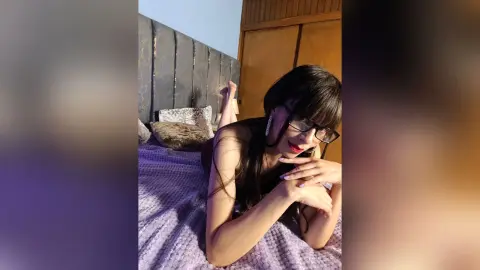 DacotaVelle wearing glasses and looking down while lying on a purple bed with her hands near her face.