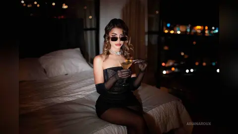 Glamorous woman in a black cocktail dress and sunglasses holding a drink in a moody, dimly lit bedroom.