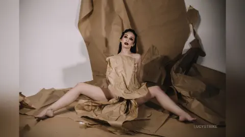 Fashion model in a creative brown paper outfit sitting amidst crumpled paper in a minimalist studio setting.
