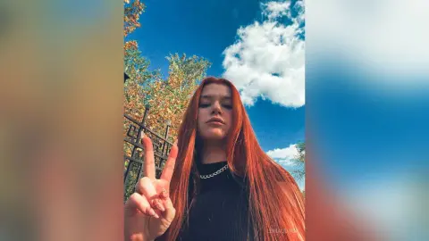 Redheaded woman in a black turtleneck making a peace sign against a bright blue sky and green trees.