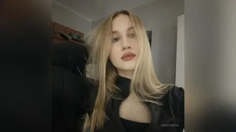 Blonde woman in a black zip-up top posing with a relaxed expression in a modern indoor setting.