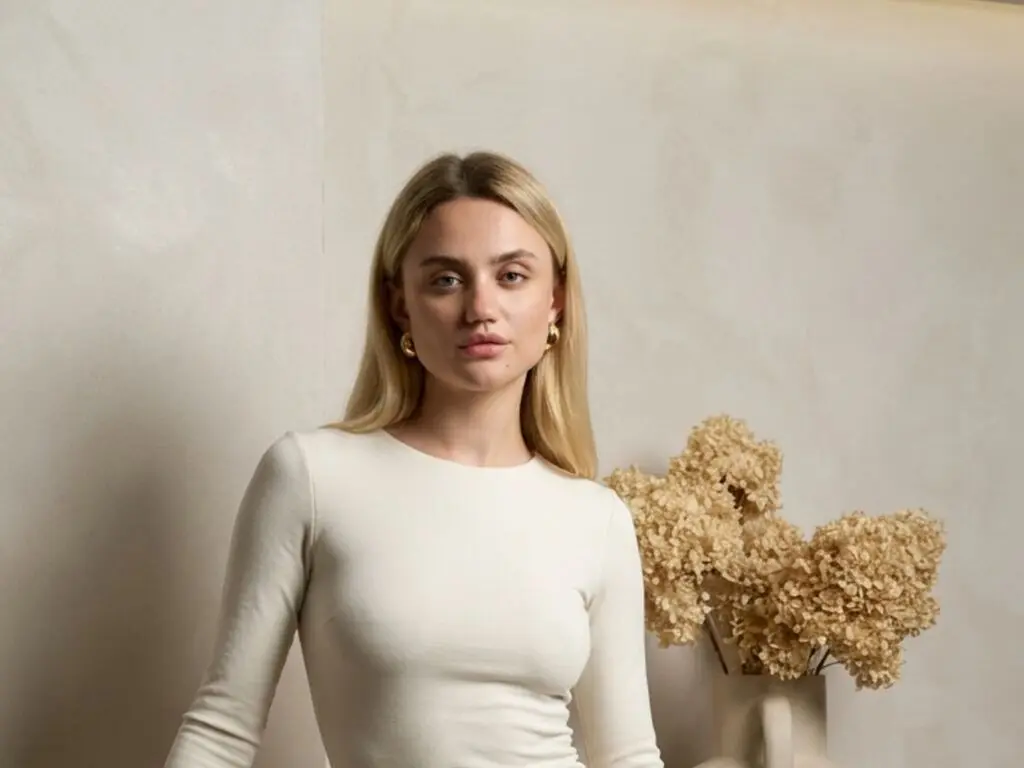 TressaGuidos poses in a white long-sleeve shirt with her blonde hair down, standing next to dried flowers against a plain wall.