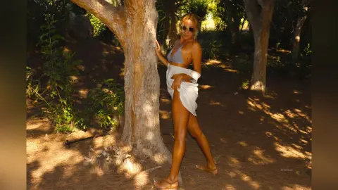 AtlamaDesmond poses outdoors by a tree, wearing a grey bikini top and a white skirt that she pulls up to expose her bare thighs and pussy.
