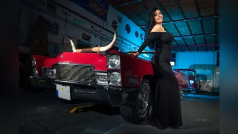 MissyJolie stands next to a red vintage car wearing a long black off-the-shoulder dress and smiling.