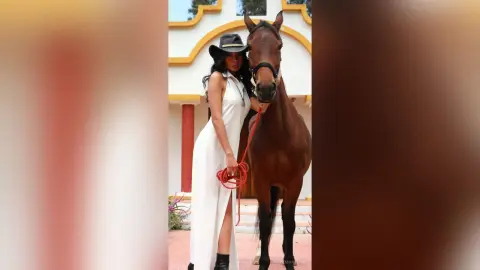 Elegant woman in a white maxi dress and black hat standing beside a brown horse in a sunny outdoor setting.