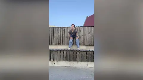 Young woman in a grey t-shirt and blue jeans posing playfully against a wooden fence under a clear blue sky.