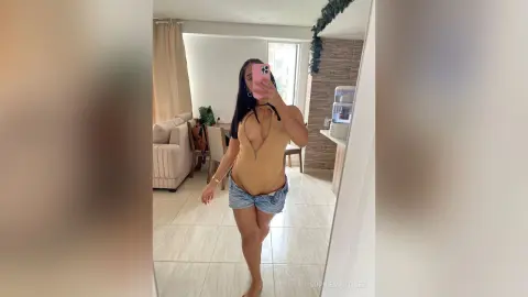 Young woman in a tan bodysuit and denim shorts taking a mirror selfie in a bright modern living room.