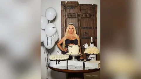 Glamorous blonde woman in a black dress celebrating with white balloons and a cake near a rustic wooden door.