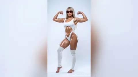 NatashaCastello poses in a white cutout bodysuit and tall white socks, flexing her muscular arms and showing off her toned body.