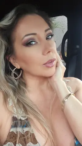 Glamorous woman with blonde hair and smoky eye makeup wearing a lace patterned top in a car.
