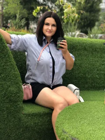 SweetEmmyGirl wearing a light blue button-down shirt and black shorts, sitting on a green grass bench while holding a black cup.