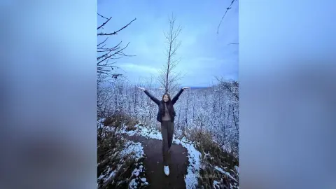 Happy woman in a black jacket raising arms on a snowy forest trail under a blue winter sky.