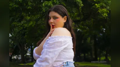 Young woman in a white off-the-shoulder blouse posing thoughtfully against a lush green forest background.