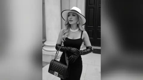 Anelise poses in a black dress and white hat, wearing long black gloves and a pearl necklace while holding a black quilted purse.