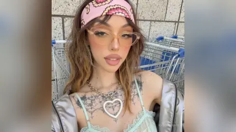 Trendy young woman with pink headband and gold glasses wearing a silver jacket and heart necklace outdoors.