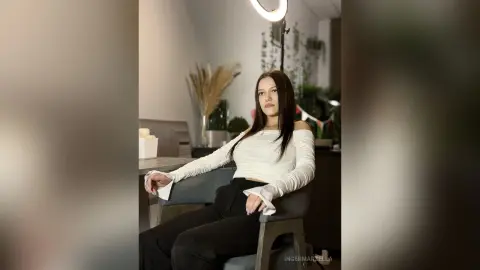 IngerMarzella sits in a chair wearing a white long-sleeved top and black pants, looking toward the camera with long dark hair.