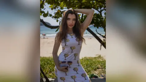 Beautiful woman in a white tree-print summer dress posing under tropical trees on a sunny beach.
