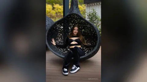 Stylish woman in black cutout top and trousers relaxing in a modern hanging egg chair outdoors.