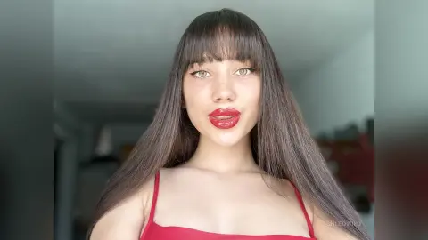 Young woman with long brown hair and red lipstick wearing a red tank top in a bright indoor setting.