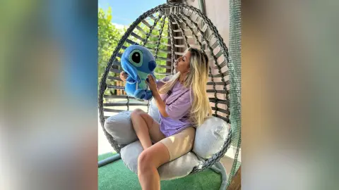 Anita sits in a hanging egg chair wearing a purple t-shirt and holding a blue Stitch plush toy.