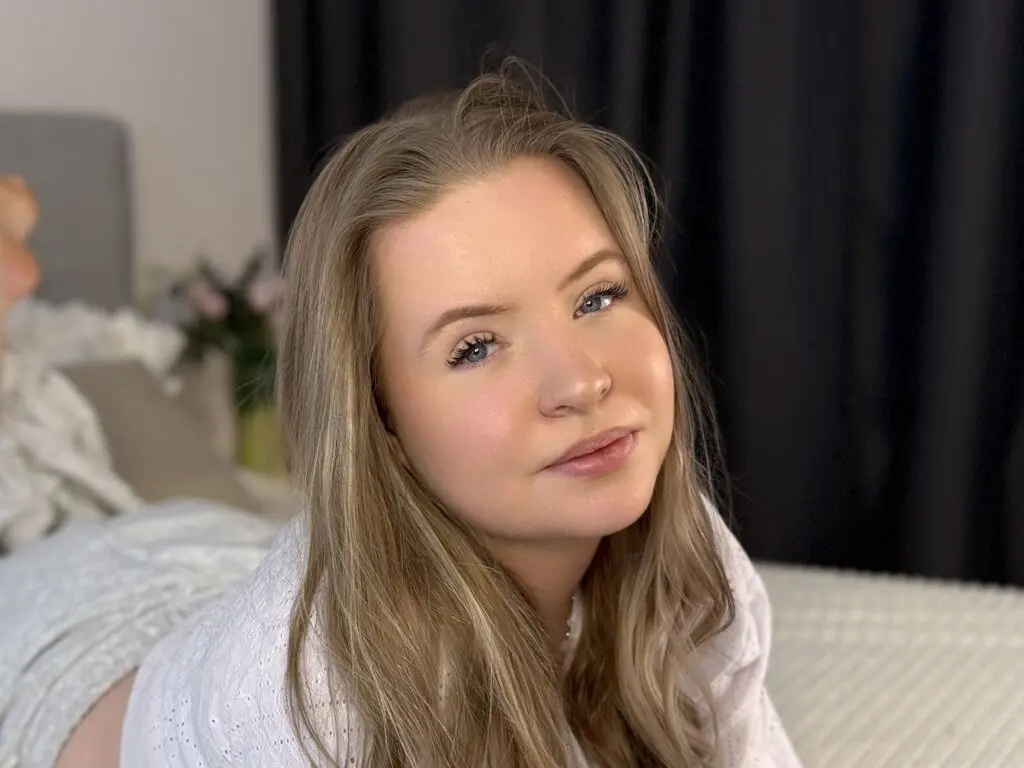 Smiling young woman with long blonde hair wearing a white blouse in a cozy, bright bedroom setting.
