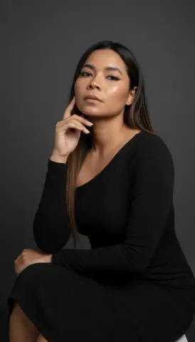 XinemaBonet poses with her hand to her face, wearing a long-sleeved black dress against a dark grey background.