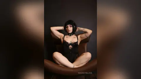 Gothic woman in black lace bodysuit sitting cross-legged in a chair against a dark moody background.