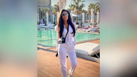 ArianaKroft stands outdoors by a swimming pool wearing white trousers and a white top with a black crossbody bag and sunglasses.