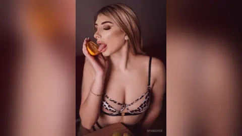 Blonde woman in leopard print bikini eating an orange slice in a warm, moody indoor setting.