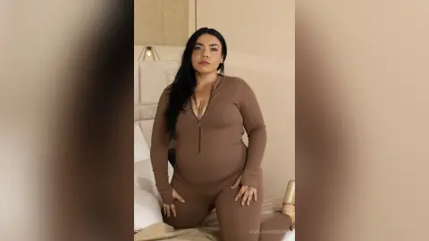 Curvy woman in a brown ribbed jumpsuit posing confidently on a bed in a cozy bedroom setting.