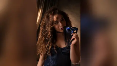 Mysterious woman with curly hair in a black lace outfit holding a dark masquerade mask in moody lighting.