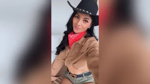 Smiling woman in a black cowboy hat, red bandana, and tan shirt posing in a bright indoor setting.