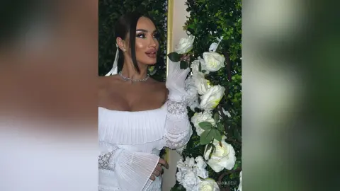 AnnaKarev poses in a white lace dress with her dark hair pulled back, standing next to a wall of white roses.