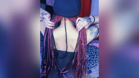 Woman with long red and black micro braids wearing a black tank top in a soft blue-lit indoor setting.
