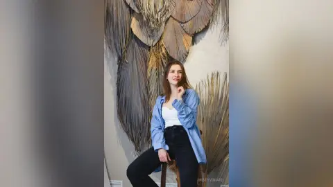 Young woman in a blue striped shirt and black jeans posing against a decorative dried palm leaf wall backdrop.