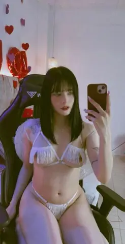 Young woman with dark hair in white lace bikini sitting in a gaming chair for a casual indoor selfie.