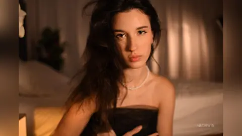Young woman with long dark hair in a black strapless dress posing in a warm, moody bedroom setting.