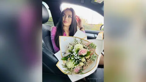 Smiling woman in a pink top holding a large bouquet of white and pink roses inside a car.